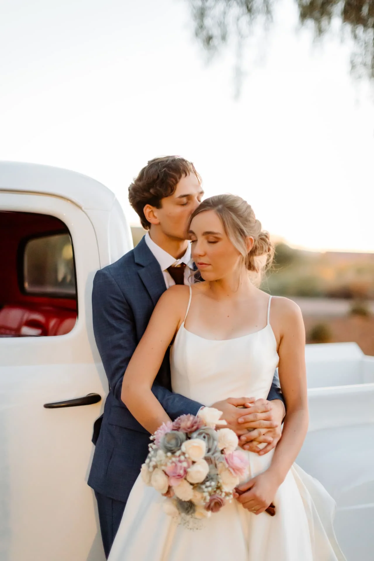 Groom kissing the bride's temple beside a vintage truck — romantic golden hour Arizona wedding portrait
