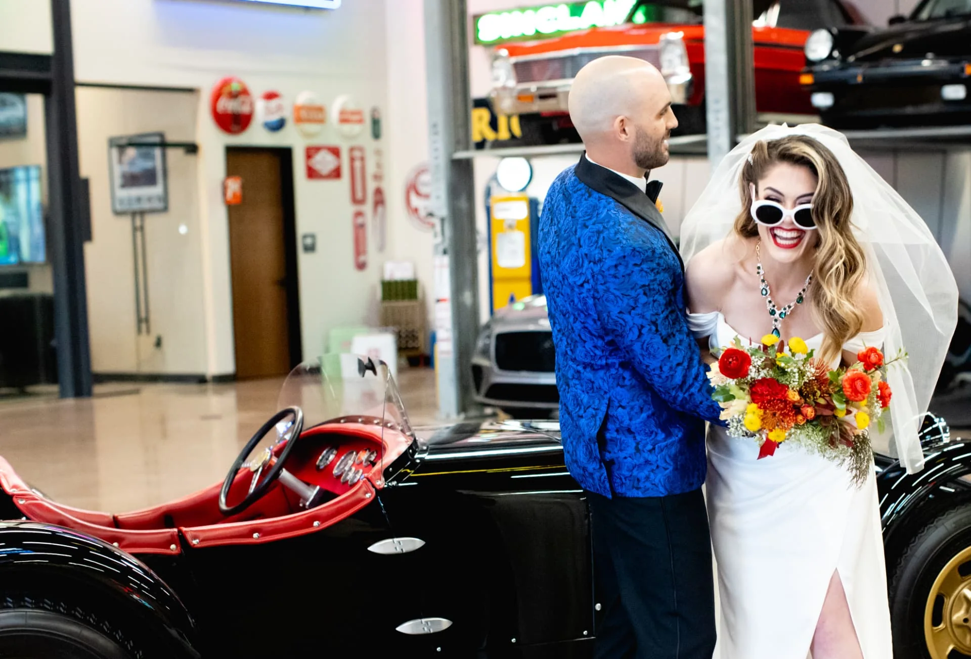 Bride in white sunglasses and a red lip laughing beside a black classic roadster while the groom in a blue jacquard jacket watches — The Vault wedding in Scottsdale, Arizona