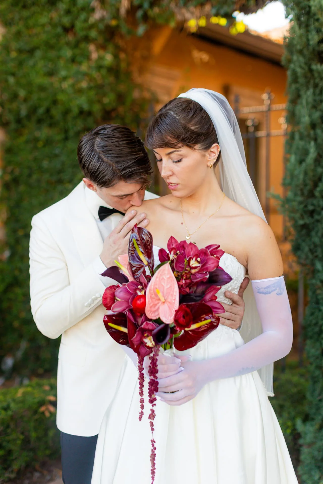 Bride holding a burgundy orchid and anthurium bouquet while the groom kisses her shoulder — romantic Scottsdale Arizona wedding