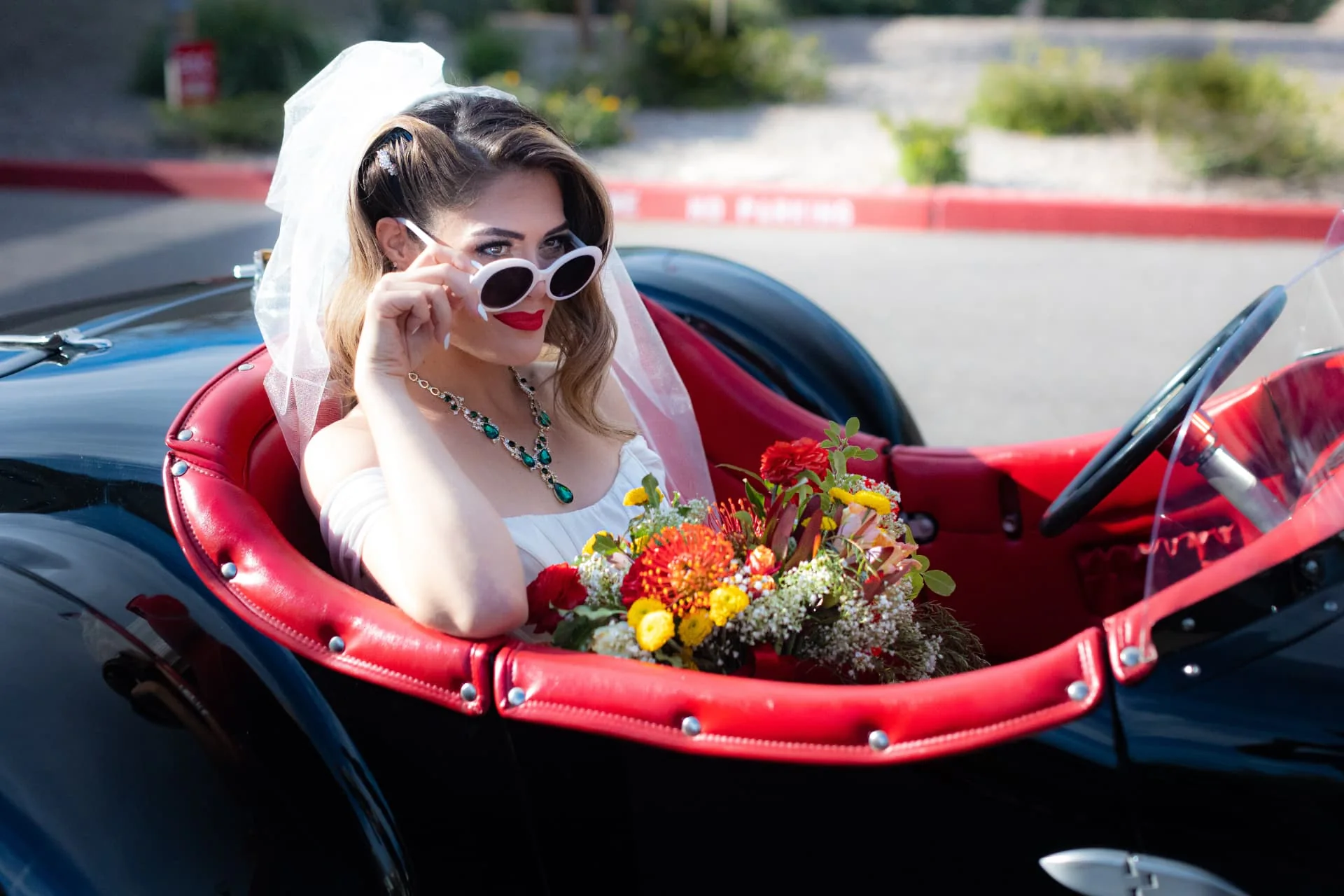 Bride in sunglasses and a red lip sitting in a black classic roadster with red leather interior — vintage car wedding at The Vault in Scottsdale, Arizona
