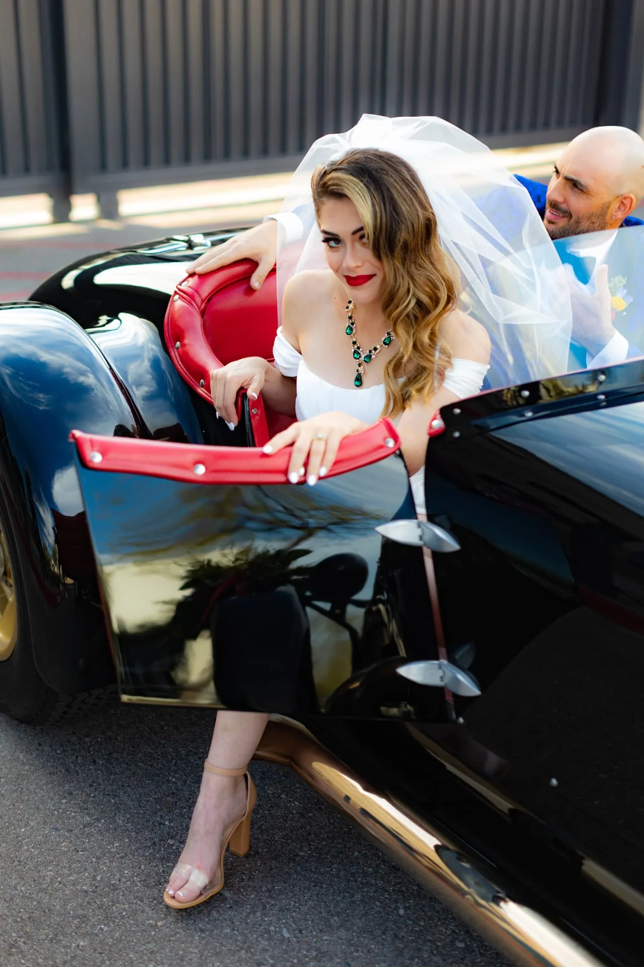 Bride stepping out of a classic black roadster while the groom in a blue floral brocade jacket watches — Scottsdale Arizona wedding photographer
