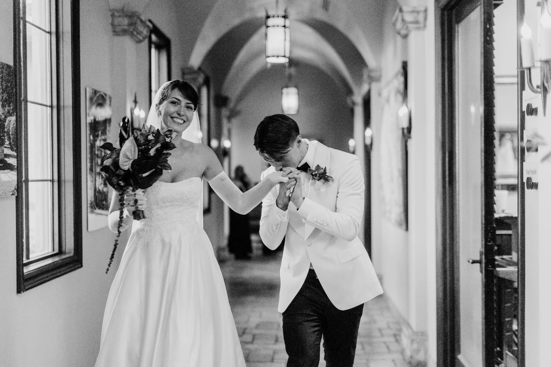 Bride in opera gloves and a groom in a cream dinner jacket walking down a vaulted hallway in black and white — romantic Scottsdale Arizona wedding